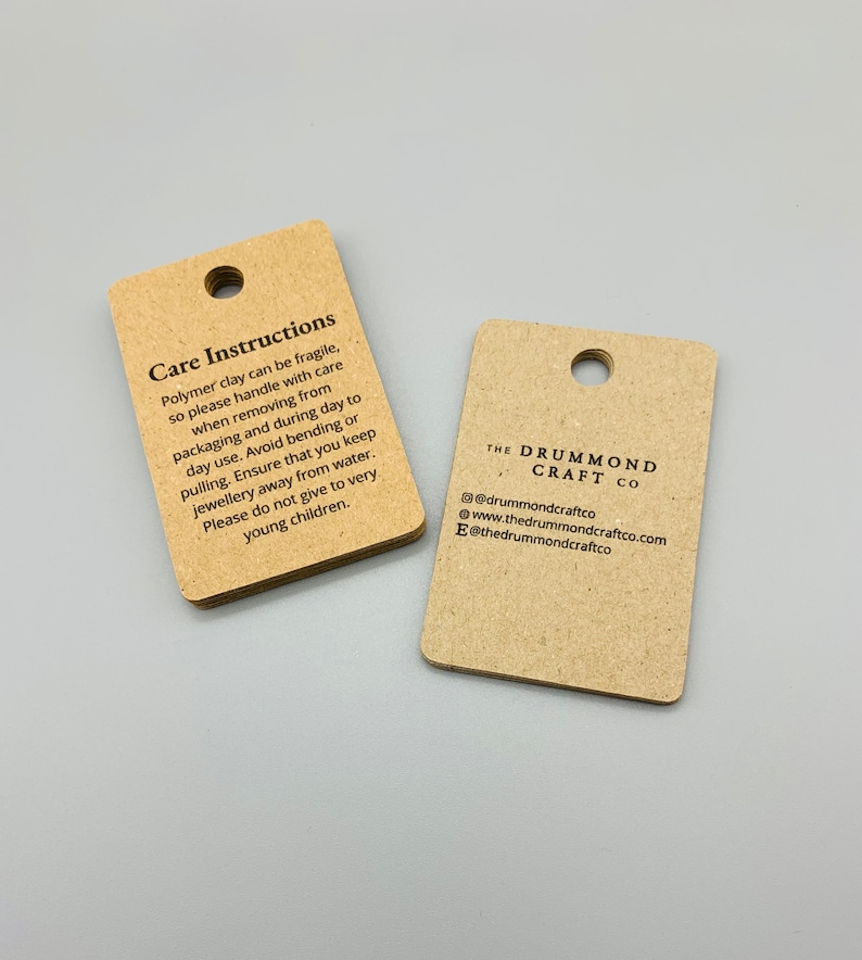 K&ouml;nnte beinhalten: Zwei braune Papieretiketten mit schwarzem Text. Das erste Etikett enth&auml;lt Pflegehinweise f&uuml;r Schmuck aus Polymer Clay. Das zweite Etikett tr&auml;gt den Text "The Drummond Craft Co." mit Kontaktinformationen.