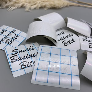 May include: Several white sheets of transfer paper with the phrase "Small Business Bitch" printed in black cursive lettering. The sheets are laid out on a gray surface.