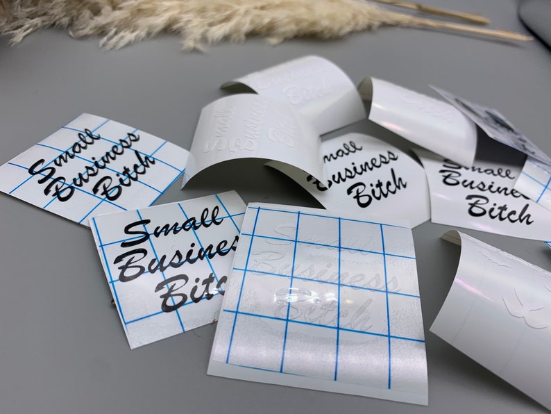 May include: Several white sheets of transfer paper with the phrase "Small Business Bitch" printed in black cursive lettering. The sheets are laid out on a gray surface.