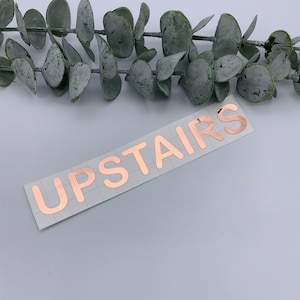 May include: Rose gold vinyl sticker with the word "UPSTAIRS" in a modern font. The sticker is on a white background.