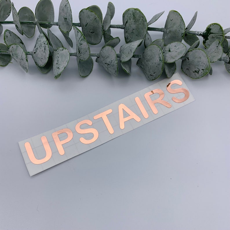May include: Rose gold vinyl sticker with the word "UPSTAIRS" in a modern font. The sticker is on a white background.