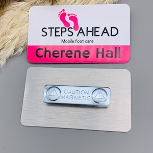 Puede incluir: Una placa de identificación blanca y rosa con el texto "STEPS AHEAD Mobile foot care" y el nombre "Chereene Hall". La placa de identificación tiene un respaldo magnético plateado con una etiqueta de advertencia que dice "CAUTION MAGNETIC".