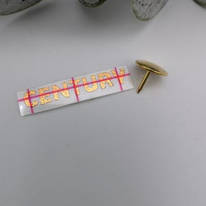 May include: A gold-colored push pin with a round head and a sharp point. The pin is on a white surface next to a white sticker with the word "CENTURY" printed in gold letters.