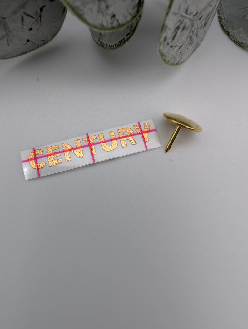 May include: A gold-colored push pin with a round head and a sharp point. The pin is on a white surface next to a white sticker with the word "CENTURY" printed in gold letters.