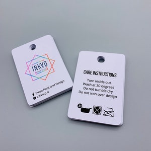 K&ouml;nnte beinhalten: Zwei wei&szlig;e rechteckige Etiketten mit abgerundeten Ecken. Das Etikett auf der linken Seite zeigt ein farbiges Logo mit dem Text "INKVO Print & Design" und Social-Media-Icons. Das Etikett auf der rechten Seite zeigt den Text "CARE INSTRUCTIONS" und Anweisungen zum Waschen und B&uuml;geln des Kleidungsst&uuml;cks. Die Anweisungen beinhalten ein Waschmaschinensymbol mit der Zahl 30, ein Trockner-Symbol mit einem Kreuz dar&uuml;ber und ein B&uuml;geleisensymbol mit einem Kreuz dar&uuml;ber.