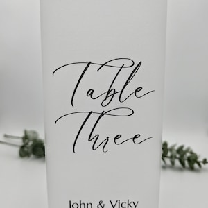 May include: A white table number card with elegant black script. The card reads "Table Three" above the names "John & Vicky" and the date "09.05.26". The card is rectangular and stands upright.