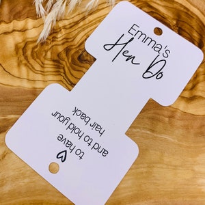 May include: White card with a cut-out shape for a favor bag. The card says "Emma's Hen Do" and "To have and to hold your hair back."