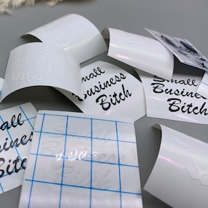 May include: White vinyl sheets with the text "Small Business Bitch" printed in black. The sheets are on a gray surface.