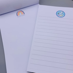 Puede incluir: Dos blocs de notas blancos con líneas y diferentes logotipos. El primer bloc de notas tiene un logotipo de arcoíris con el texto "I'm in Stitches Handmade Goods". El segundo bloc de notas tiene un círculo azul con una paloma blanca y el texto "Dove's Ink".