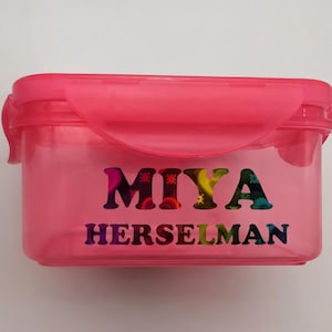 May include: A rectangular, translucent pink plastic food storage container with a matching snap-lock lid. The front of the container features the name MIYA HERSELMAN printed in a bold, multi-colored, patterned font with various circular designs.
