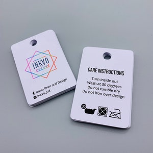 K&ouml;nnte beinhalten: Zwei wei&szlig;e rechteckige Anh&auml;nger mit abgerundeten Ecken. Der Anh&auml;nger auf der linken Seite hat ein farbenfrohes Logo mit dem Text "INKVO Print & Design" und Social-Media-Icons. Der Anh&auml;nger auf der rechten Seite hat den Text "PFLEGEANWEISUNGEN" und Anweisungen, das Kleidungsst&uuml;ck auf links zu drehen, bei 30 Grad zu waschen, nicht im Trockner zu trocknen und nicht &uuml;ber das Design zu b&uuml;geln. Der Anh&auml;nger enth&auml;lt au&szlig;erdem drei Symbole: eine Waschmaschine mit der Zahl 30 darin, ein durchgestrichener Trockner und ein durchgestrichenes B&uuml;geleisen.