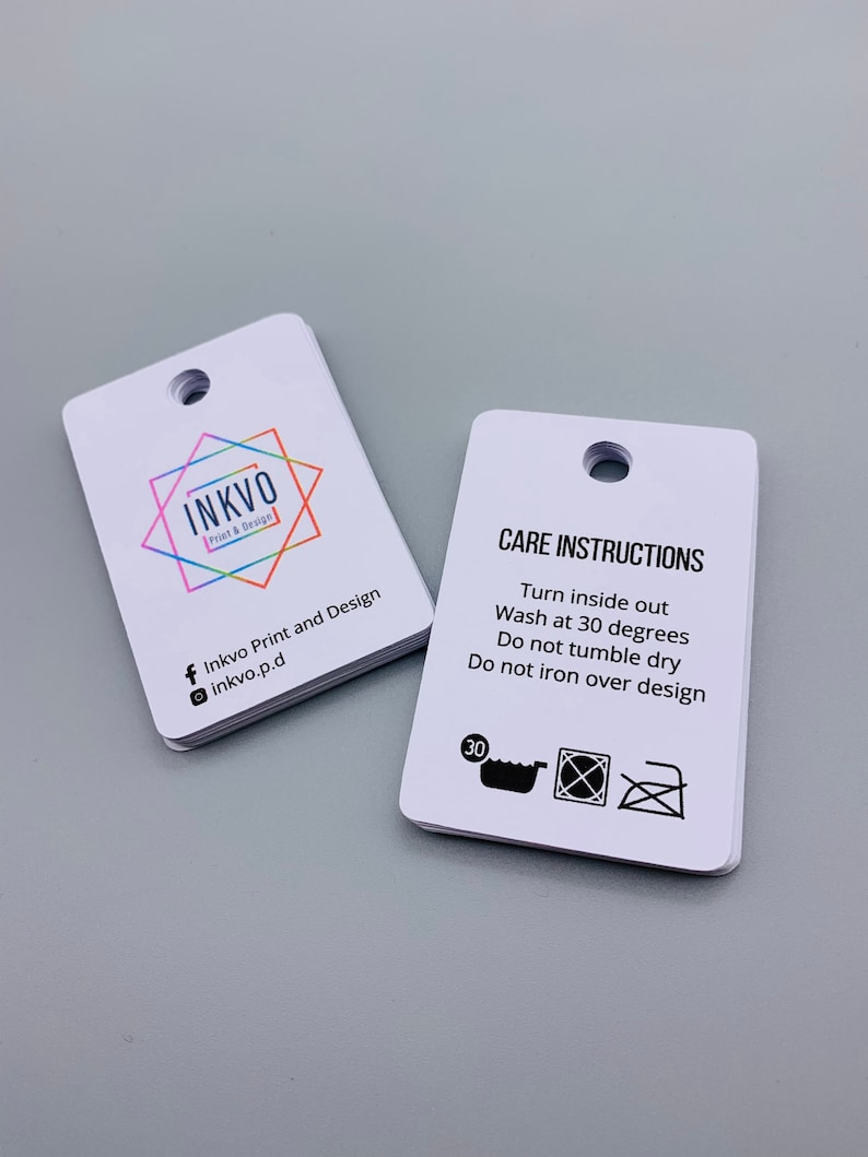 K&ouml;nnte beinhalten: Zwei wei&szlig;e rechteckige Anh&auml;nger mit abgerundeten Ecken. Der Anh&auml;nger auf der linken Seite hat ein farbenfrohes Logo mit dem Text "INKVO Print & Design" und Social-Media-Icons. Der Anh&auml;nger auf der rechten Seite hat den Text "PFLEGEANWEISUNGEN" und Anweisungen, das Kleidungsst&uuml;ck auf links zu drehen, bei 30 Grad zu waschen, nicht im Trockner zu trocknen und nicht &uuml;ber das Design zu b&uuml;geln. Der Anh&auml;nger enth&auml;lt au&szlig;erdem drei Symbole: eine Waschmaschine mit der Zahl 30 darin, ein durchgestrichener Trockner und ein durchgestrichenes B&uuml;geleisen.