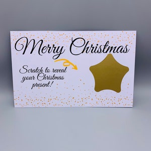 Puede incluir: Una tarjeta navideña blanca para rascar con confeti dorado y un área para rascar dorada en forma de estrella. La tarjeta dice "Feliz Navidad" en una elegante escritura, con el texto "¡Raspa para revelar tu regalo de Navidad!"