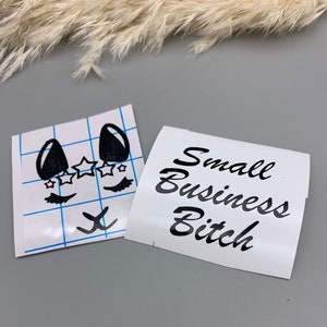May include: Two white vinyl stickers with black lettering and designs. One sticker features a cartoon face with eyelashes, stars, and a small 'x' below. The other sticker says "Small Business Bitch".