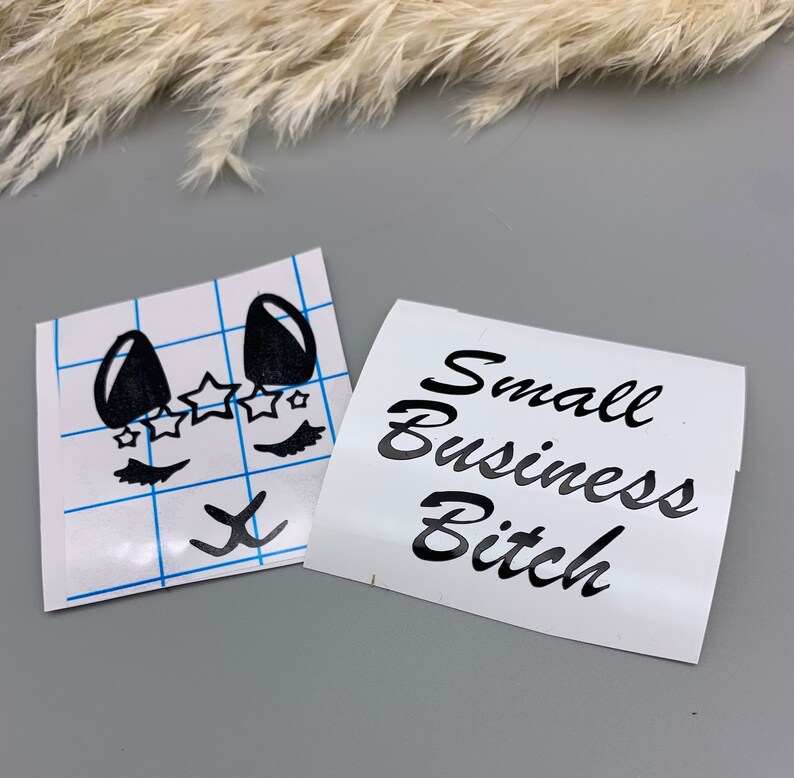 May include: Two white vinyl stickers with black lettering and designs. One sticker features a cartoon face with eyelashes, stars, and a small 'x' below. The other sticker says "Small Business Bitch".