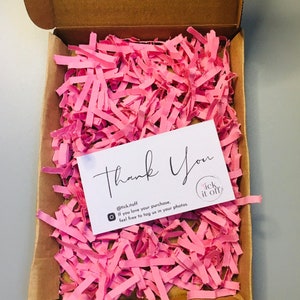 Puede incluir: Una tarjeta de agradecimiento blanca con texto negro que dice "Gracias" y "@tick.itoff Si te encanta tu compra, no dudes en etiquetarnos en tus fotos." La tarjeta está sobre papel de seda rosa dentro de una caja de cartón marrón.
