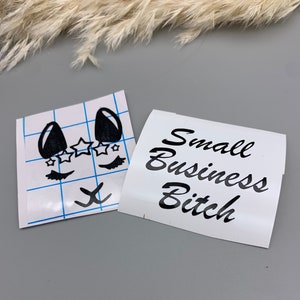 May include: Two white sheets of paper with black vinyl lettering. One sheet has a cartoon face with eyelashes, stars, and a nose. The other sheet has the text "Small Business Bitch".