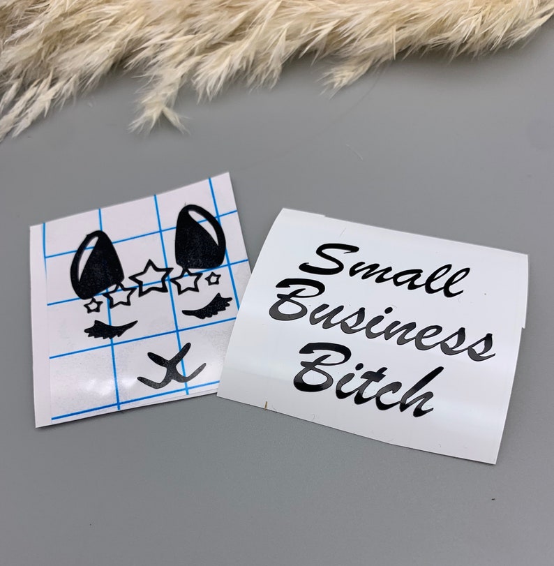 May include: Two white sheets of paper with black vinyl lettering. One sheet has a cartoon face with eyelashes, stars, and a nose. The other sheet has the text "Small Business Bitch".
