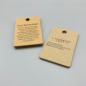 K&ouml;nnte beinhalten: Zwei braune Papieretiketten mit Pflegehinweisen und Kontaktinformationen f&uuml;r The Drummond Craft Co. Das erste Etikett tr&auml;gt den Text "Care Instructions" und beschreibt, wie man mit Polymer Clay Schmuck umgeht. Das zweite Etikett enth&auml;lt den Firmennamen und die Kontaktinformationen, einschlie&szlig;lich der Website und der Social-Media-Handles.
