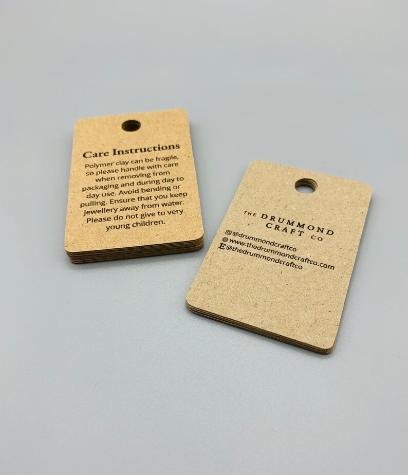K&ouml;nnte beinhalten: Zwei braune Papieretiketten mit Pflegehinweisen und Kontaktinformationen f&uuml;r The Drummond Craft Co. Das erste Etikett tr&auml;gt den Text "Care Instructions" und beschreibt, wie man mit Polymer Clay Schmuck umgeht. Das zweite Etikett enth&auml;lt den Firmennamen und die Kontaktinformationen, einschlie&szlig;lich der Website und der Social-Media-Handles.