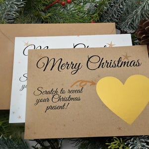 Peut inclure: Cartes de Noël avec un thème festif. La carte de devant est de couleur beige et porte l'inscription "Merry Christmas" et une zone à gratter en forme de cœur jaune. Le texte "Scratch to reveal your Christmas present!" est également visible.