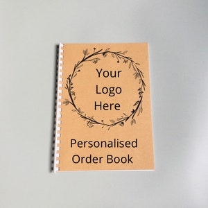Puede incluir: Un libro de pedidos de papel marrón con una encuadernación en espiral blanca. La portada presenta una corona floral negra con el texto "Your Logo Here" y las palabras "Personalised Order Book".
