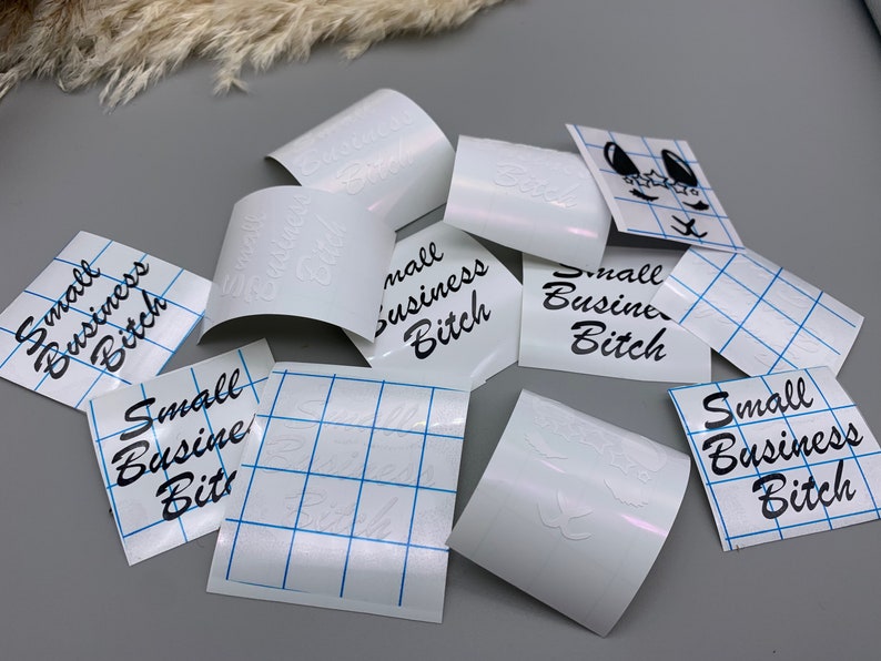 May include: A collection of white vinyl stickers with the text "Small Business Bitch" in black lettering. The stickers are on a white gridded backing.