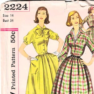 May include: Vintage Simplicity sewing pattern 2224 for a dress. The pattern is for size 14 with a bust measurement of 34 inches. The pattern shows two women wearing dresses, one in a yellow solid colour and the other in a plaid pattern.