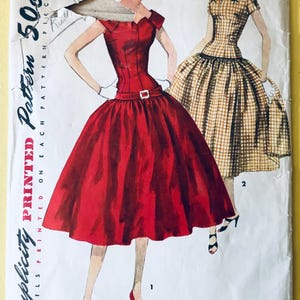 Puede incluir: Sobre de patrón Simplicity vintage con ilustraciones de dos vestidos. Uno es un vestido rojo con corpiño ajustado y falda amplia, el otro es un vestido a cuadros. El sobre está etiquetado con el número "1113" y la talla "12".