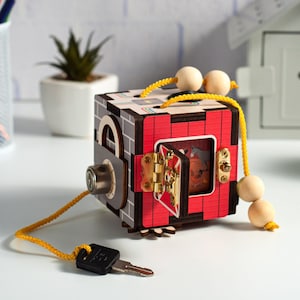 May include: A wooden toy cube with a red, gray, and brown exterior. The cube has a small door that opens and closes with a key. The door has a picture of a farm scene with animals. The cube is attached to a yellow string with beads.