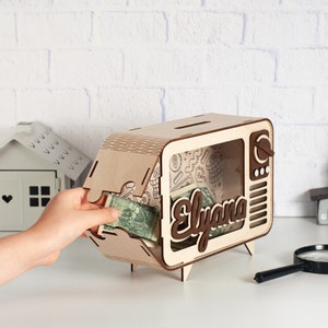 May include: A light-colored wooden piggy bank shaped like a vintage television set with a clear front panel. The name Elyana is cut out in dark wood letters on the front. The interior features a patterned background with money inside the box.