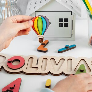 May include: A wooden name puzzle with the name "OLIVIA" spelled out in large letters. Each letter has a cutout shape that a child can place a corresponding wooden piece into. The puzzle includes a small, colourful hot air balloon.