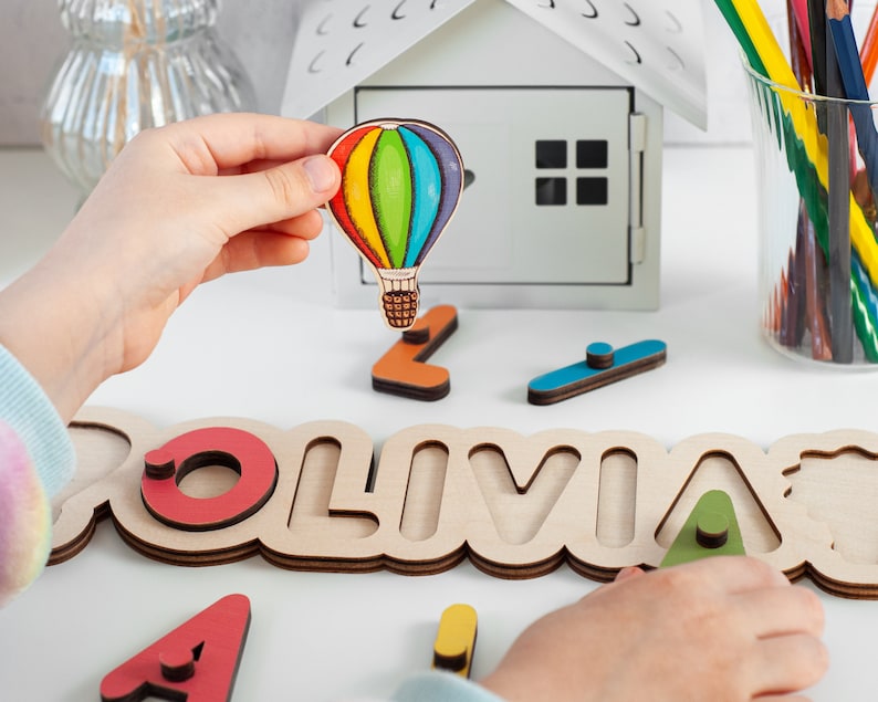 May include: A wooden name puzzle with the name "OLIVIA" spelled out in large letters. Each letter has a cutout shape that a child can place a corresponding wooden piece into. The puzzle includes a small, colourful hot air balloon.