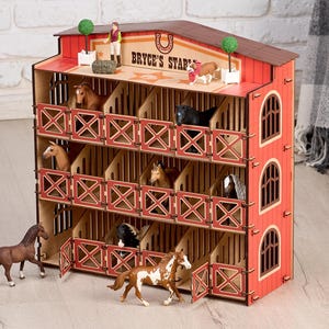 May include: A red and tan wooden horse stable with multiple stalls, each housing a toy horse. The stable has the text "Bryce's Stable" on the roof. Several toy horses are positioned in front of the stable.