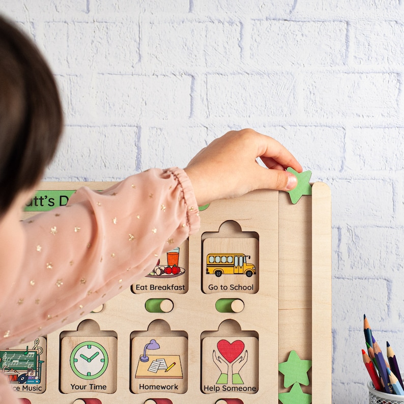 K&ouml;nnte beinhalten: Ein Holz-Aktivit&auml;tsbrett mit bunten Illustrationen und Text. Das Brett hat Schlitze zum Einsetzen von gr&uuml;nen sternf&ouml;rmigen Teilen. Der Text auf dem Brett lautet "Eat Breakfast", "Go to School", "Your Time", "Homework", "Help Someone" und "Make Music".
