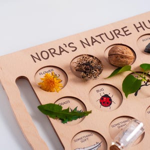May include: Wooden nature hunt board with the text "NORA'S NATURE HUNT". The board has circular cutouts containing a yellow flower, grass, a walnut, and a ladybug illustration. A small glass bottle is also visible.
