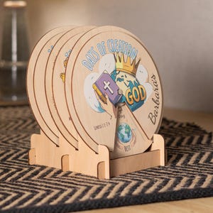 Puede incluir: Un conjunto de placas circulares de madera en un soporte de madera. La placa frontal presenta el texto Days of Creation y God, una ilustración de un globo terráqueo con una corona, una mano sosteniendo un libro con una cruz y el texto Barbara's.