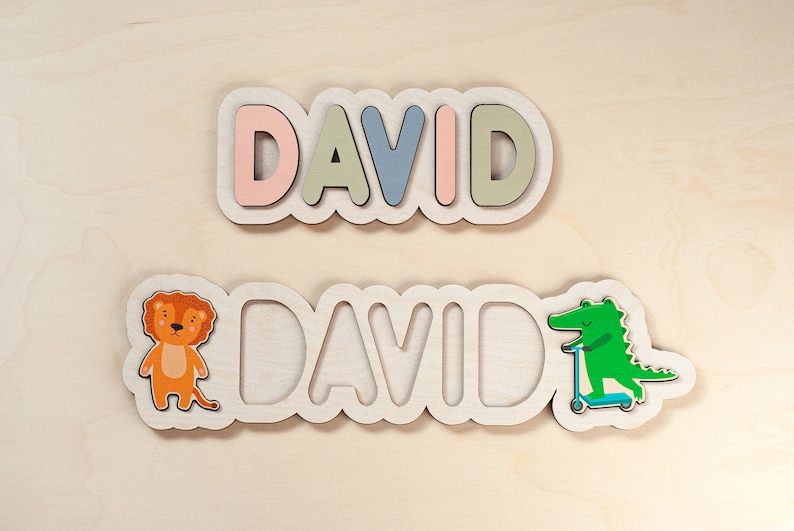 May include: Two wooden name plaques with the name "DAVID" spelled out in colourful letters. The top plaque has pastel pink, green, and blue letters. The bottom plaque has a lion and a crocodile on either side of the name.