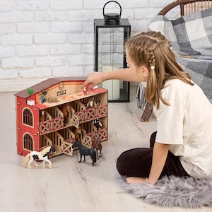 Puede incluir: Un establo de juguete de madera de dos pisos con exterior rojo, ventanas arqueadas y techo marrón. La estructura cuenta con varios compartimentos que contienen pequeñas figuras de caballos de plástico, con la palabra STABLE y un icono de herradura.