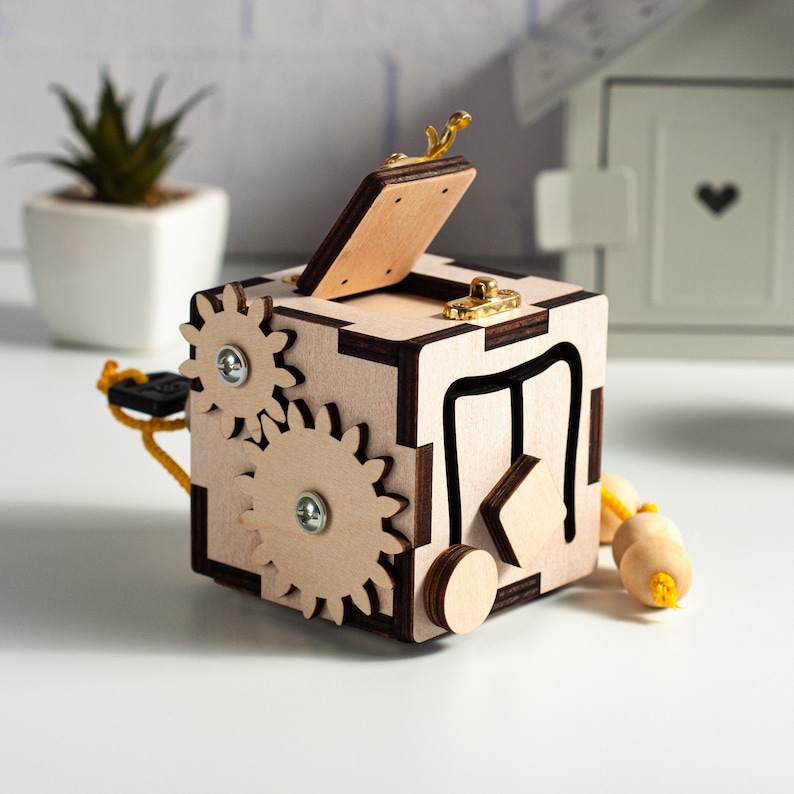 May include: A wooden toy box with a hinged lid, featuring gears, a sliding door, and a small latch. The box is painted in a light brown color with dark brown accents.