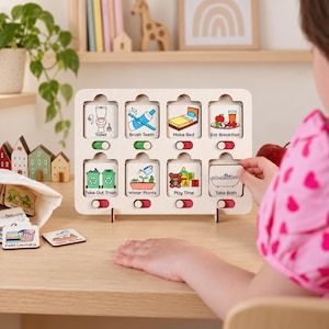Puede incluir: Un tablero de tareas de madera clara con ocho ranuras y fichas cuadradas extra&iacute;bles. Cada ficha muestra un icono colorido y texto para tareas diarias como Toilet, Brush Teeth, Make Bed, Eat Breakfast, Take Out Trash, Water Plants, Play Time y Take Bath. Cada ranura tiene un interruptor deslizante.