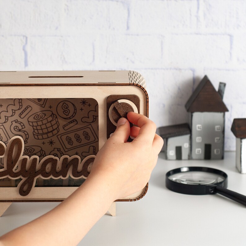 May include: A rectangular wooden piggy bank shaped like a vintage television set with a light wood frame, a dark wood dial, and a transparent front panel featuring engraved dollar signs and coin graphics. The name Elyana is displayed in dark wood.