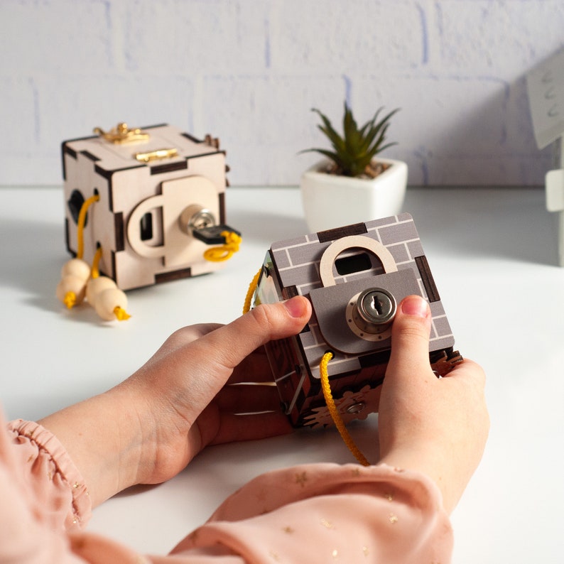 May include: A child plays with two wooden toy boxes. One box is brown with a lock and key, the other is light brown with a keyhole. Both boxes have yellow string attached.