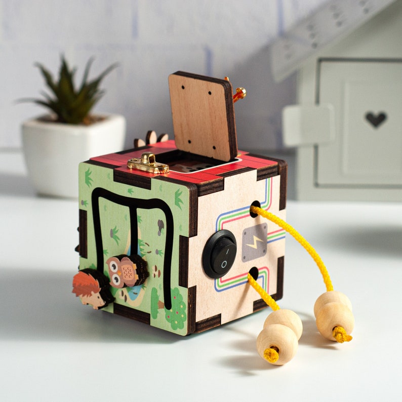 May include: A wooden toy cube with a colorful design and various activities, including a sliding door, a maze, and a switch. The cube has a yellow cord with wooden beads attached.