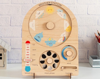 May include: A wooden weather station board featuring a rotating dial with weather icons, a sliding temperature gauge, a moon phase wheel, a wind speed dial, and a humidity slider. The text "WEATHER STATION" is printed at the top of the board.