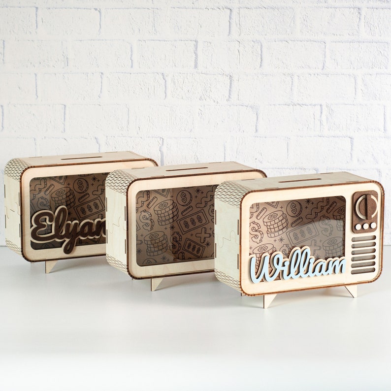 May include: Three rectangular wooden piggy banks shaped like vintage televisions. Each features a clear front panel, a top coin slot, and a patterned interior with dollar signs and coins. Two banks display the names Elyana and William in script.