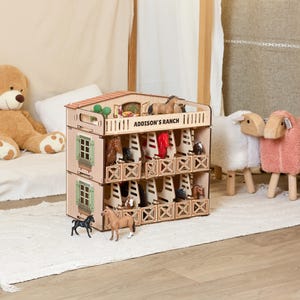 May include: A two-storey wooden toy horse stable with the text "ADDISON'S RANCH" printed on the top level. The structure features multiple stalls, green window shutters, and includes several small plastic horse figurines placed inside.
