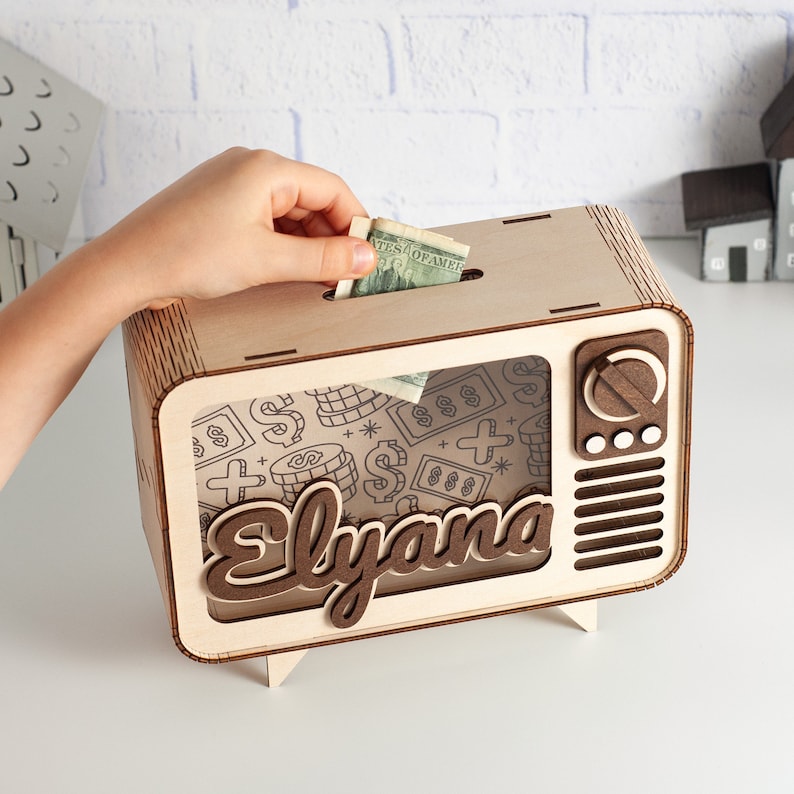 May include: A wooden piggy bank shaped like a vintage television set with a light wood frame and dark wood accents. The front features the name Elyana in dark wood script, a speaker grille, and a dial. The screen displays a pattern of dollar signs.