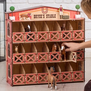 May include: A wooden toy horse stable with multiple stalls, painted red and natural wood. The sign reads "BRYCE'S STABLE". The stable is decorated with toy horses and figures. The toy has a decorative horseshoe on top.