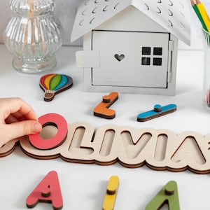 May include: Wooden name puzzle featuring the name Olivia in large letters. The puzzle pieces are colourful and have a variety of shapes. The puzzle is on a white surface with other wooden toys.